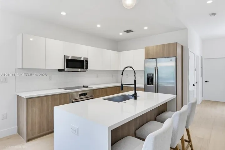 Kitchen featuring flat-panel two-tone cabinetry, stone-surface countertops, a center island with an integrated sink and gooseneck faucet, stainless steel appliances, and wood-finish flooring