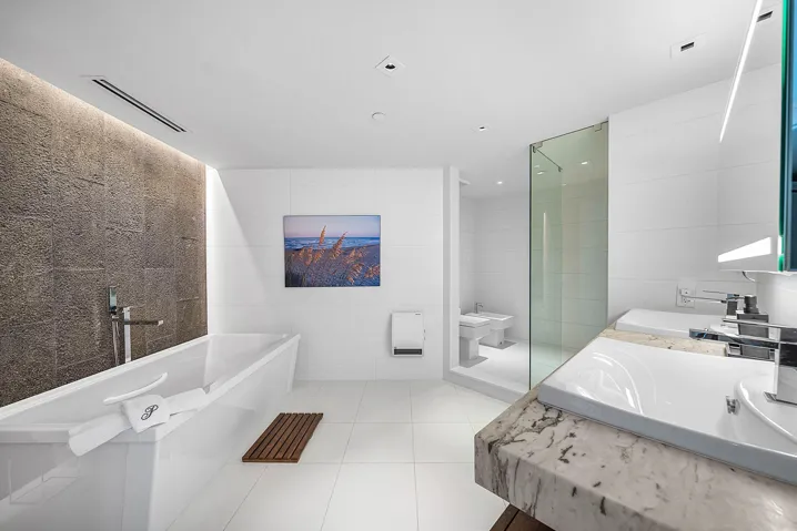 Bathroom featuring a freestanding white soaking tub, textured stone accent wall, dual-sink vanity with stone countertop, frameless glass-enclosed shower, and large-format tile flooring