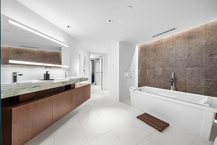 Floating wood-finish double vanity with stone countertop, large LED-backlit mirror, soaking tub, textured stone feature wall, and recessed ceiling lighting