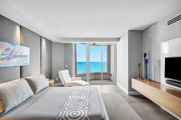Floor-to-ceiling glass sliding doors opening to a balcony, wall-to-wall upholstered headboard paneling, wall-mounted wood-finish media console, and large-format tile​​‌​​​​‌​​‌‌​‌‌​​​‌‌​‌​‌​‌​​​‌​​ flooring