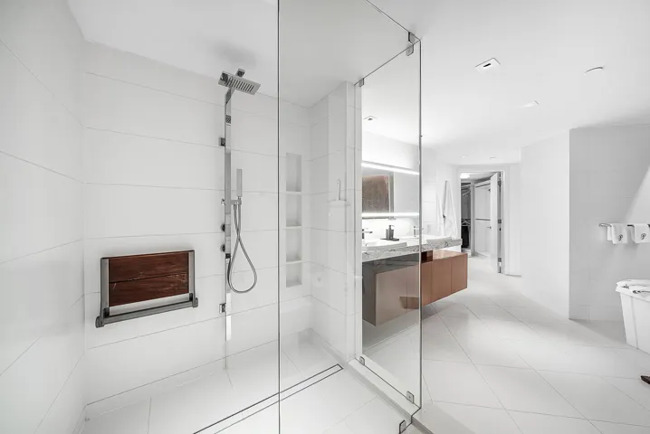 Walk-in shower with floor-to-ceiling tile, frameless glass enclosure, wall-mounted shower panel with handheld sprayer, built-in shelving, and integrated wooden fold-down seat