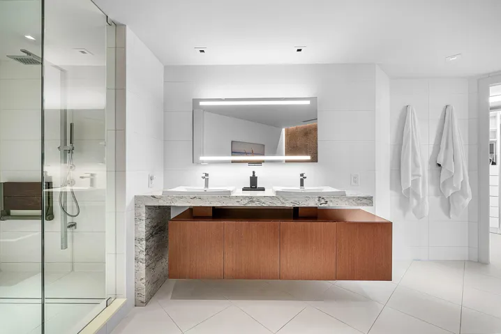 Floating wood-finish vanity with dual integrated sinks and stone-surfaced waterfall countertop, complemented by an illuminated vanity mirror and glass-enclosed walk-in shower