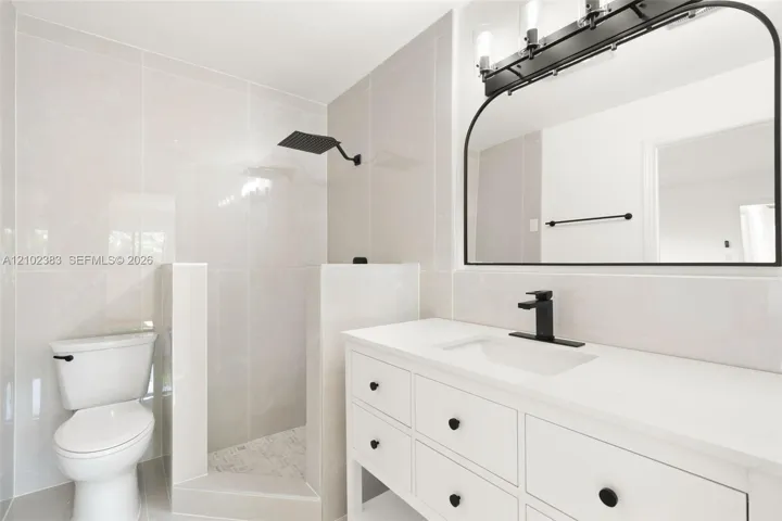 Primary bathroom featuring a white vanity with a quartz countertop, matte black faucet, wall-mounted rain shower head, arched vanity mirror, and light-toned wall tile