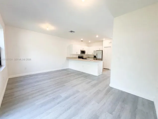 Open-concept interior featuring light wood-finish flooring, white shaker-style cabinetry, stainless steel appliances, and a breakfast bar with stone-surface countertop