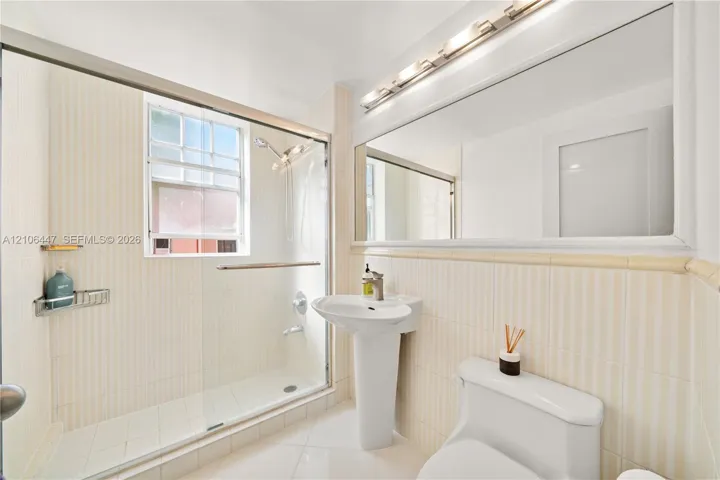Full-width vanity mirror with integrated multi-bulb lighting, pedestal sink, glass-enclosed walk-in shower with tiled surround, and ceramic tile flooring