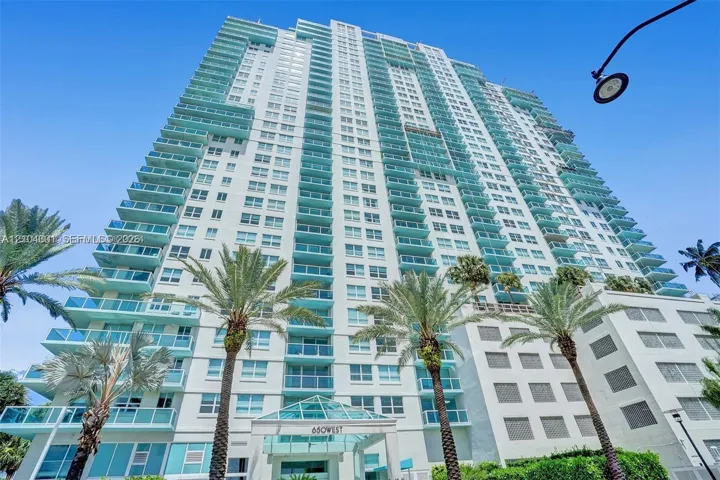 High-rise residential exterior featuring a light-toned facade, multiple levels of private glass-railed balconies, and a geometric glass-paneled entrance canopy, accented by mature palm trees