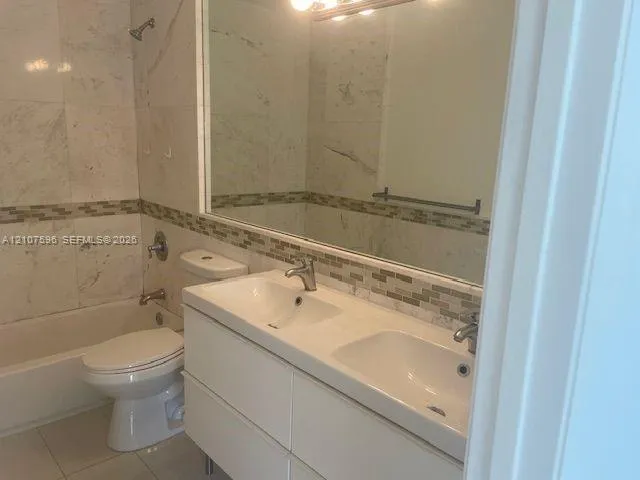 Bathroom featuring a dual-sink vanity with a white countertop, large wall-mounted mirror, mosaic tile backsplash, and a tiled shower-tub combination