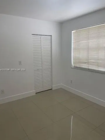 Neutral-toned tile flooring, white walls, and a closet with white louvered bi-fold doors, complemented by a window with installed horizontal blinds