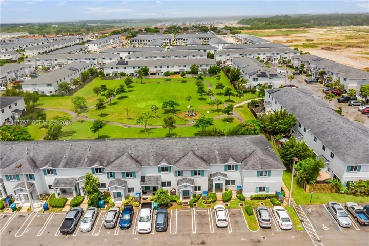 Aerial perspective of a multi-unit residential complex featuring light-toned siding, dark shingle roofs, designated surface parking, and extensive landscaped green space with paved pedestrian walkways