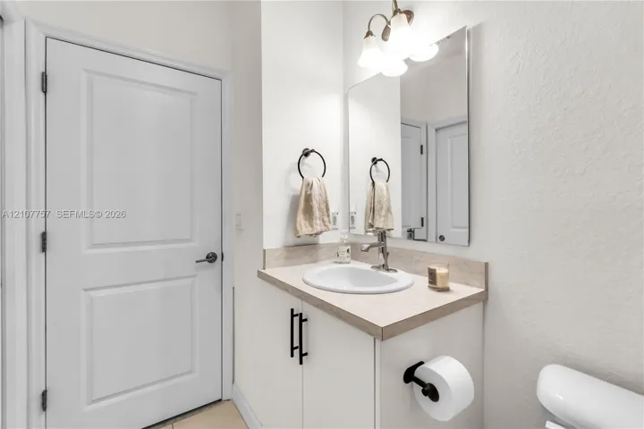 Vanity featuring a light-toned countertop, undermount oval sink, matte black hardware, and an attached three-light fixture above a rectangular mirror