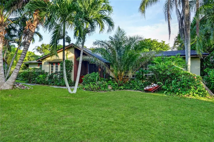 Single-story exterior featuring a low-pitch roofline, painted siding, mature palm trees, and extensive tropical landscaping