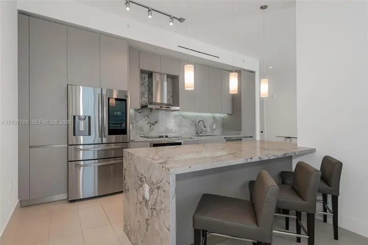 Contemporary kitchen featuring flat-panel cabinetry, stone countertops with waterfall edge, stainless steel appliances, integrated range hood, and pendant lighting