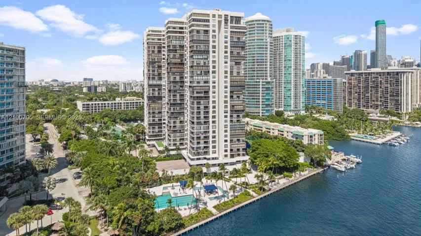 High-rise residential building featuring floor-to-ceiling glass paneling, a ground-level swimming pool, expansive waterfront frontage, and mature landscaping