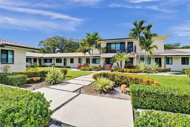 Stucco exterior building featuring a paved walkway, mature palm trees, manicured hedges, and a second-level balcony with metal railing