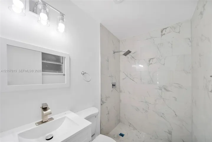 Bathroom featuring large-format stone-patterned tile shower surround, integrated sink vanity, wall-mounted light fixture, and metallic finish faucet hardware