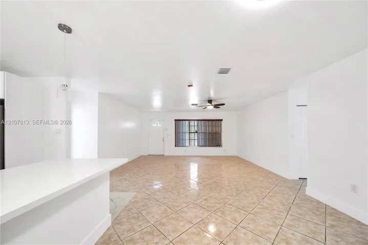 Open-concept interior featuring light-toned tile flooring, white cabinetry, a built-in kitchen island, a ceiling fan, and a single-light pendant fixture