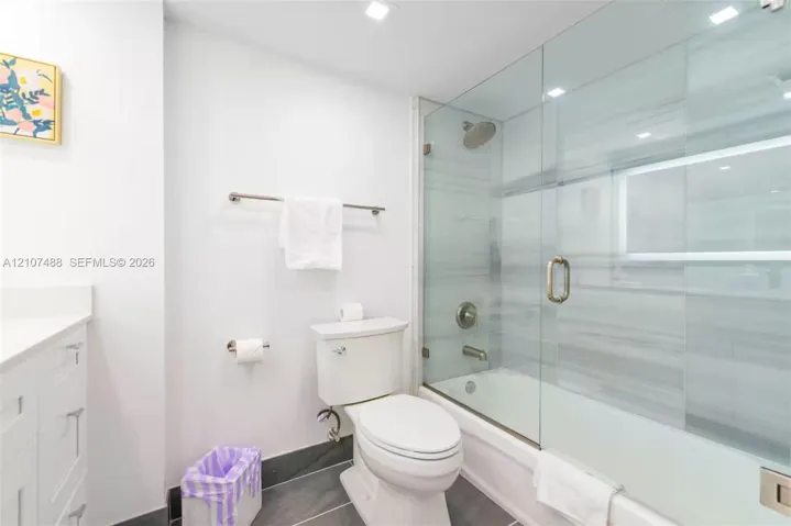 Bathroom featuring a tub-shower combination enclosed by a frameless glass door, horizontal tile wall surround, recessed lighting, white vanity cabinet with a stone countertop, and gray large-format tile flooring