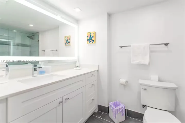 Bathroom featuring a vanity with white cabinetry and a solid-surface countertop, an integrated rectangular sink, a large wall-mounted LED-backlit mirror, and dark tile flooring