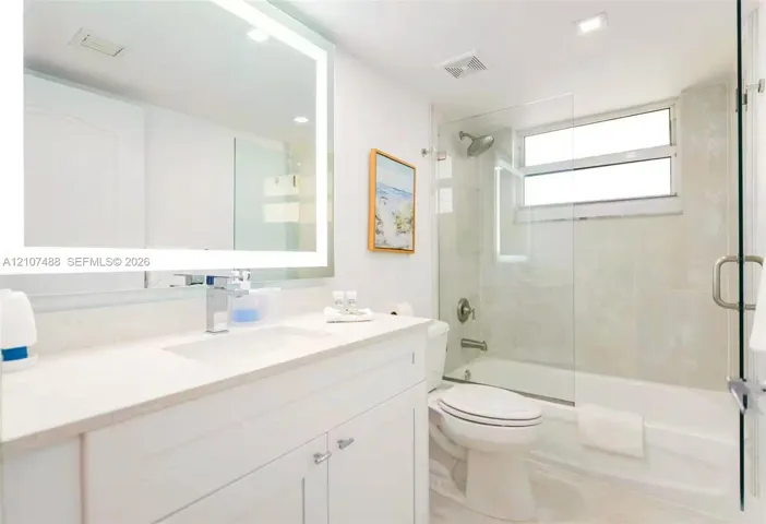 Bathroom featuring a white vanity cabinet with an integrated sink, backlit vanity mirror, glass-enclosed shower and tub combination with tile surround, and a horizontal window