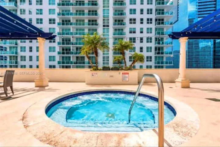 In-ground spa featuring a stone-style coping, metal handrail, dual blue-tinted pergolas supported by architectural columns, and built-in planters with palm trees