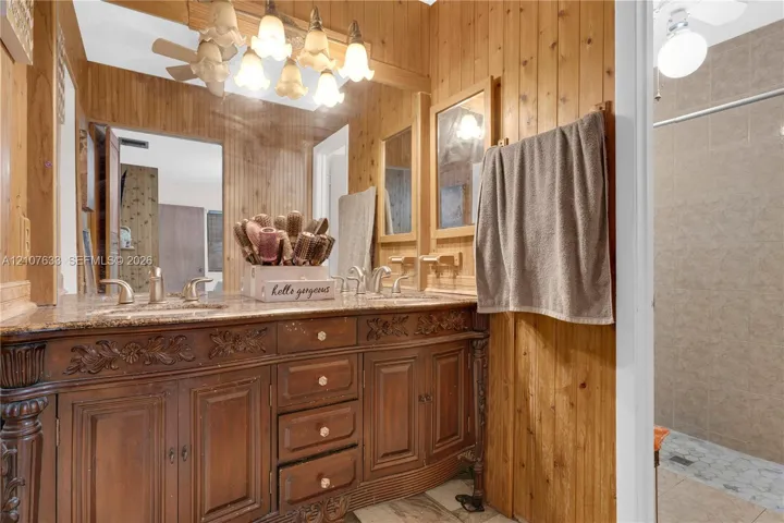 Dual-sink vanity with ornate wooden cabinetry and stone countertop, wood-paneled walls, and multi-bulb decorative light fixture