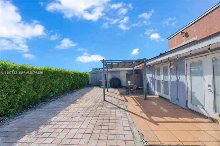 Paved outdoor patio featuring a metal pergola, dual glass-paneled exterior doors, and a natural perimeter hedge