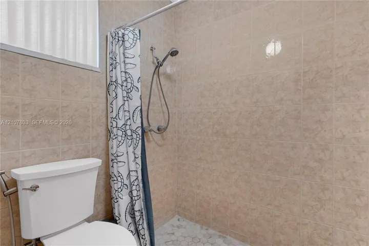 Shower stall featuring floor-to-ceiling tile, a handheld shower fixture, a white ceramic toilet, and a light-toned hexagonal tile floor