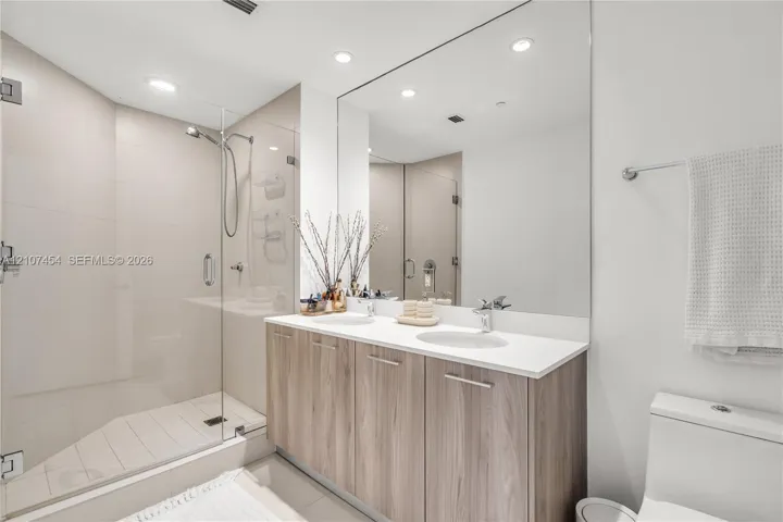 Double vanity featuring wood-finish cabinetry, solid surface countertops, dual undermount sinks, and a frameless glass-enclosed shower with tiled walls