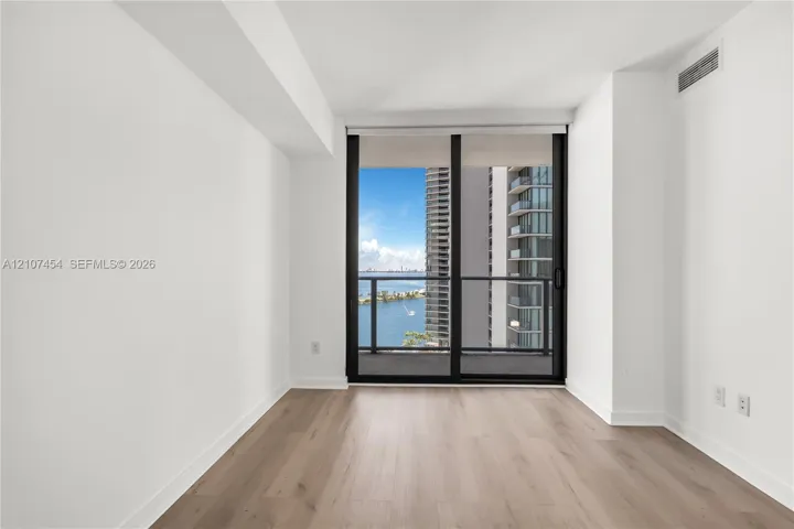 Floor-to-ceiling sliding glass doors opening to a private balcony with water and urban scenery, complemented by light wood-finish flooring