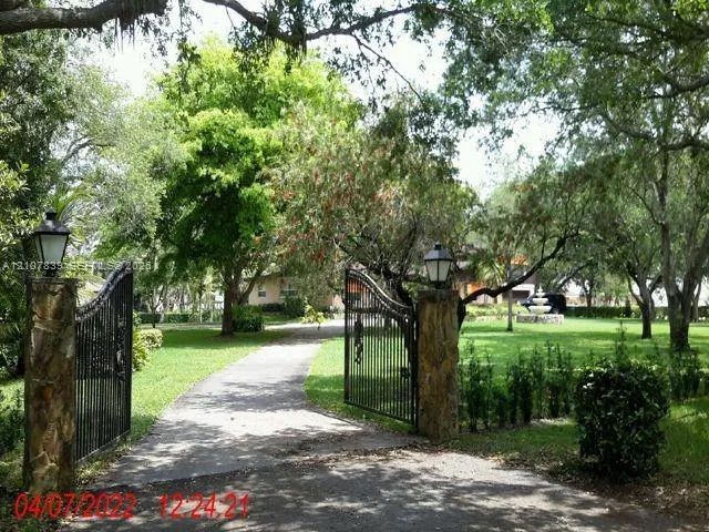 Gated entrance featuring stone masonry pillars, wrought iron fencing, mounted exterior lighting fixtures, and a landscaped grounds area with mature trees and a fountain