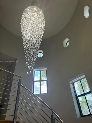 Tapered multi-light crystal chandelier suspended from a vaulted ceiling, featuring circular and rectangular windows and a metal-railing staircase