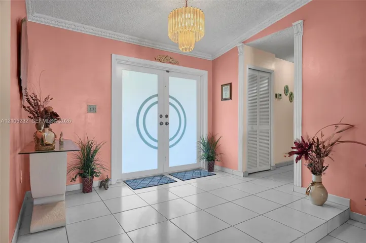 Entryway featuring double doors with frosted glass inserts, tile flooring, tiered crystal chandelier, crown molding, and louvered closet doors