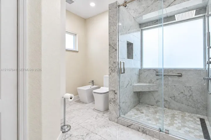 Walk-in shower with floor-to-ceiling stone-finish tile, glass enclosure, built-in bench, and hexagonal floor tile, accompanied by a bidet and dual-flush toilet