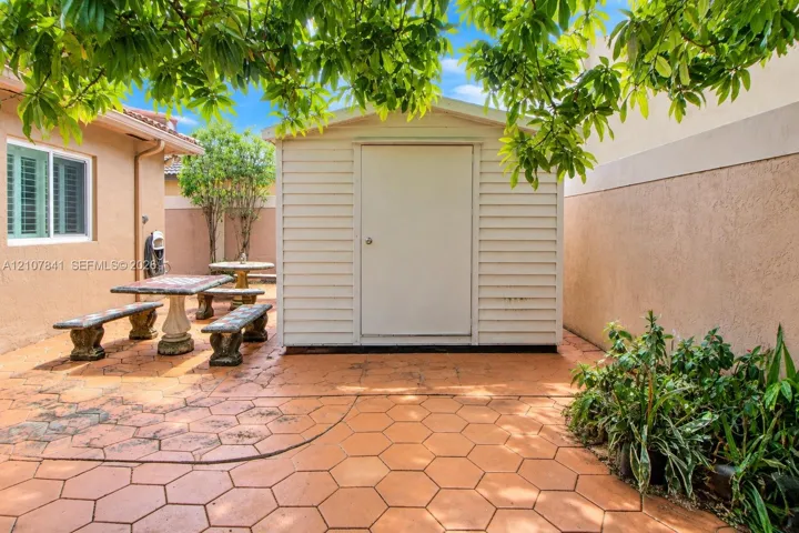 Exterior space featuring a neutral-toned storage shed, hexagonal paver hardscaping, and a stucco-finished wall