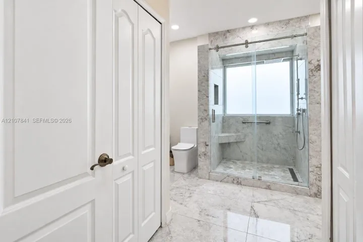 Walk-in shower with stone-finish tile surround, integrated bench seat, and sliding glass enclosure, complemented by large-format floor tiling and white dual-panel doors