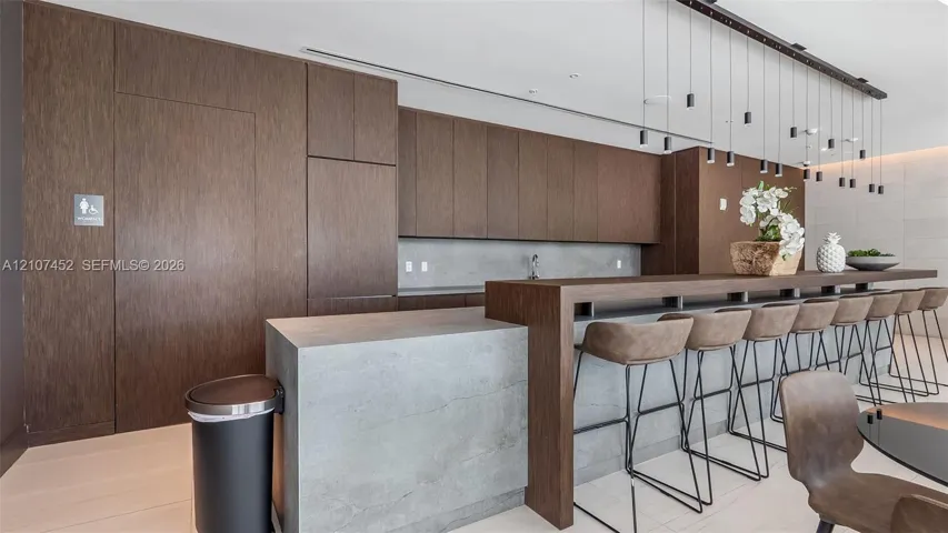 Modern kitchen featuring dark wood-finish cabinetry, gray stone-surface waterfall island with an integrated breakfast bar, and minimalist pendant lighting
