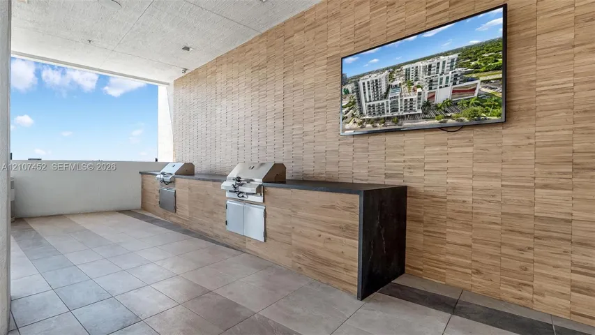 Outdoor kitchen featuring stainless steel built-in grills, custom cabinetry with wood-finish paneling, dark stone countertops, and a wall-mounted flat-screen television