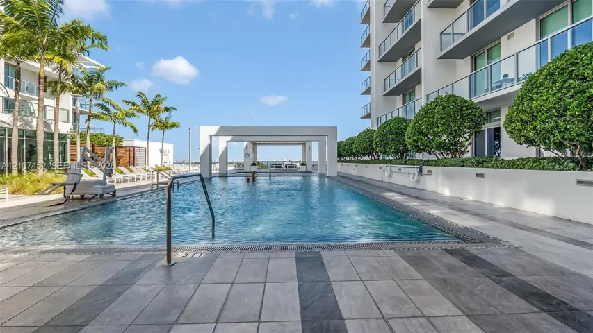 Outdoor swimming pool featuring a tiled deck, structured white pergola, perimeter landscaping, and metal railings