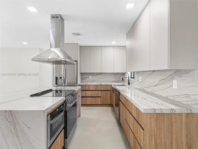 Modern kitchen featuring flat-panel wood-finish base cabinetry, light-toned upper cabinets, stone-surfaced countertops and backsplash, stainless steel range with integrated hood, and matte black plumbing fixtures