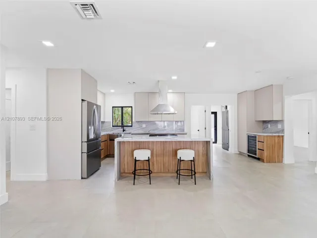 Kitchen featuring a central island with wood-finish cabinetry, stone countertops, stainless steel appliances, a range hood, and large-format tile flooring