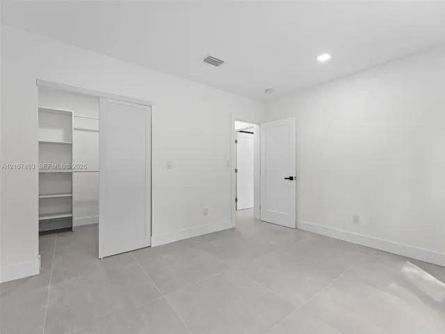 Interior space featuring large-format grey tile flooring, white walls, recessed ceiling lighting, a walk-in closet with built-in shelving, and a solid-core door with matte black hardware