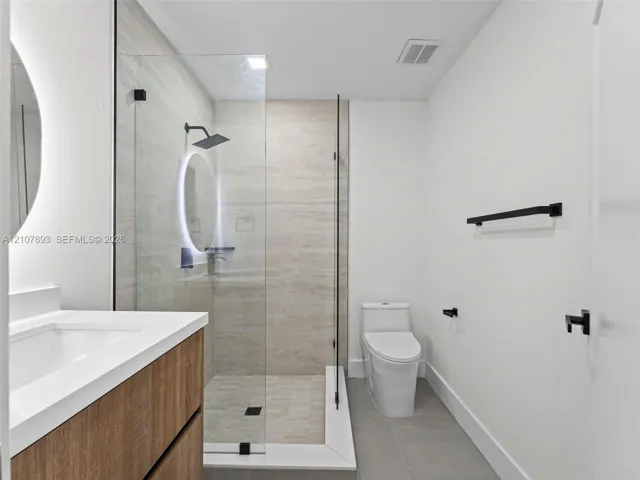 Wood-finish vanity with a rectangular sink, walk-in shower with a frameless glass enclosure, neutral tile surround, matte black rainfall shower head, and an illuminated circular mirror