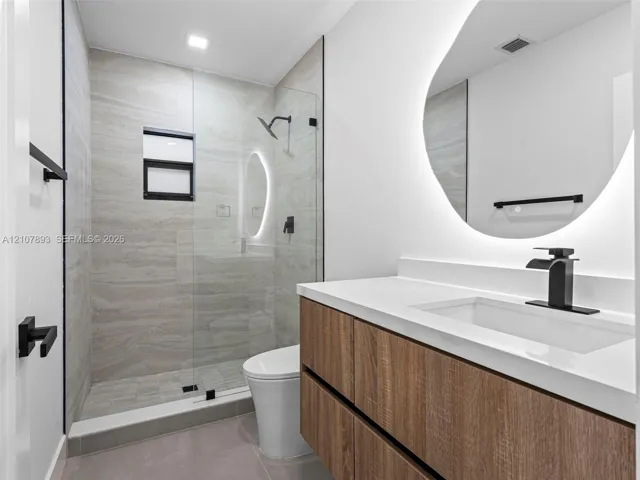 Wood-finish floating vanity with a white countertop, integrated rectangular sink, and matte black faucet, featuring a wall-mounted back-lit mirror, glass-enclosed shower with horizontal-patterned tile surround, and matte black hardware