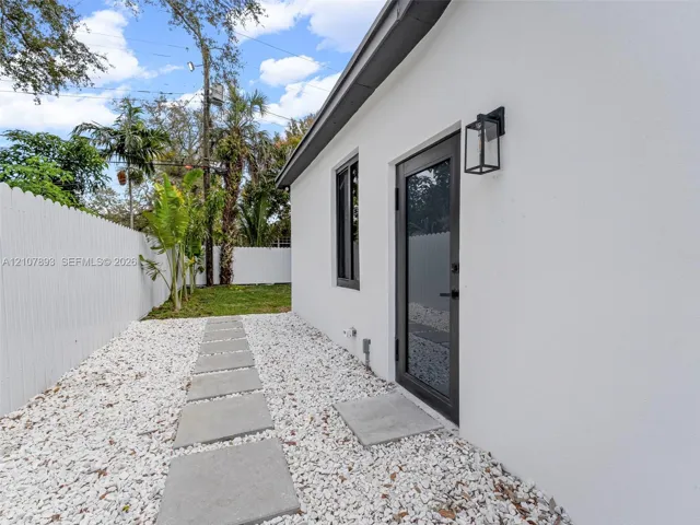 Stucco exterior featuring a dark-framed glass door, exterior wall-mounted lantern lighting, white privacy fencing, and a stone-paver walkway set in gravel