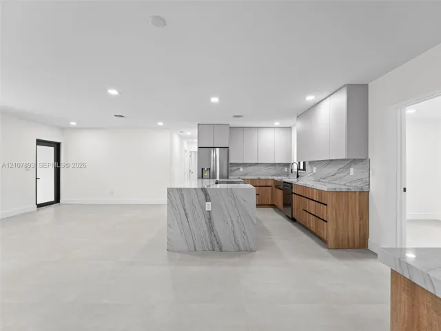 Modern kitchen featuring a central island with a stone waterfall countertop, wood-finish cabinetry, matte light gray upper cabinets, and a matching stone backsplash