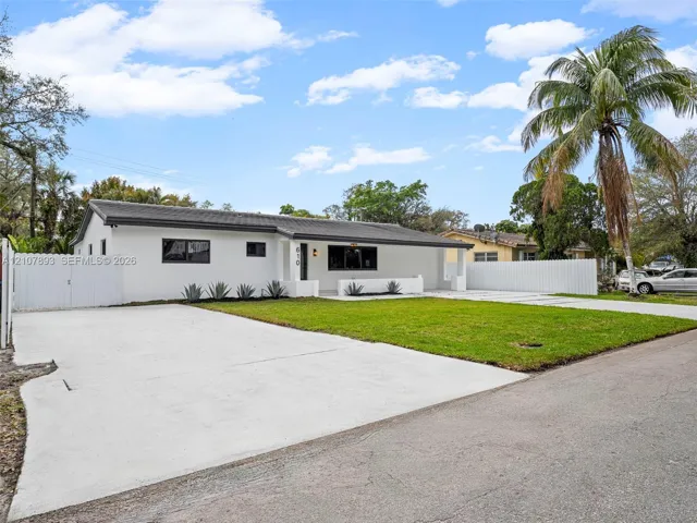 Single-story exterior featuring white stucco finish, dark-toned hip roof, expansive concrete driveway, manicured lawn, and white perimeter fencing