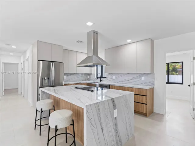 Kitchen featuring a waterfall-edge island with stone-finish countertop, dual-tone cabinetry, stainless steel range hood, and integrated cooktop