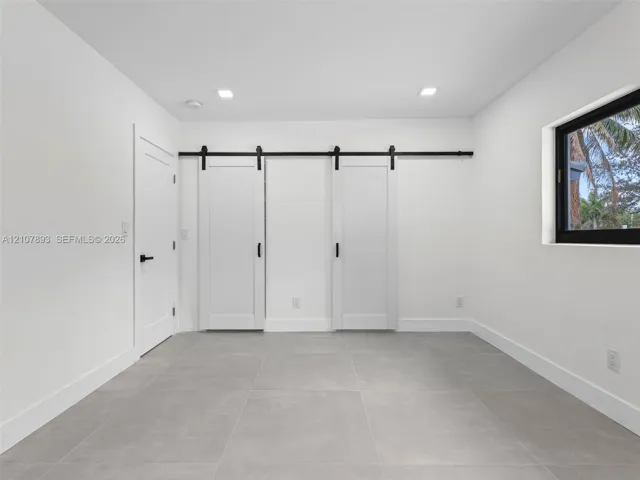 Interior room featuring large-format ceramic tile flooring, dual white sliding barn doors with matte black hardware, recessed ceiling lighting, and a window with a black frame