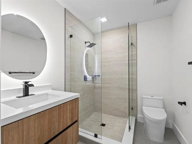 Floating vanity with wood-finish cabinetry, quartz countertop, illuminated circular mirror, glass-enclosed walk-in shower with matte black fixtures, and dual-flush toilet