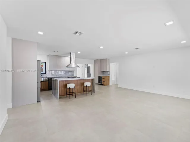 Open-concept kitchen featuring an oversized island with stone-finish waterfall edges, stainless steel range hood, built-in wine cooler, flat-panel cabinetry, and large-format tile flooring